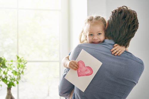 A little girl holding a card smiles while hugging her dad.