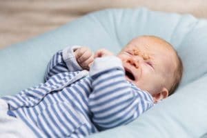 Crying infant wearing grey and blue clothes, lying on the bed.