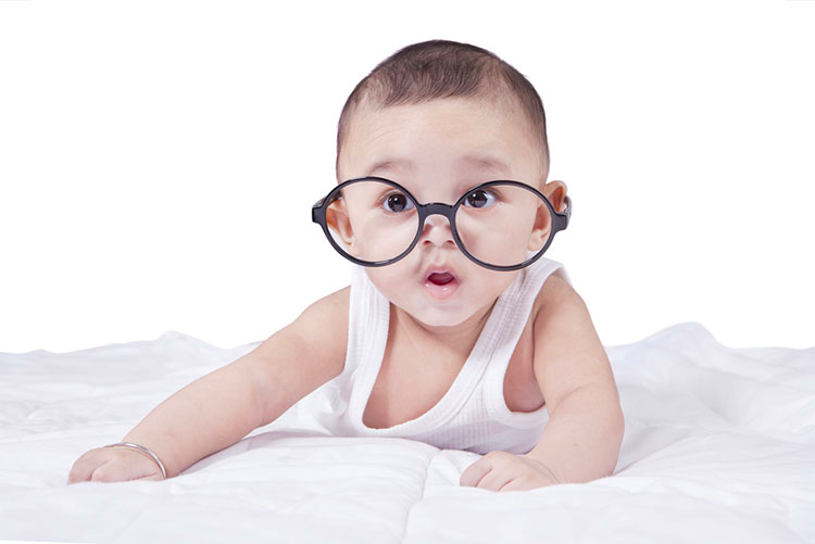 Adorable baby boy wearing big glasses, lying on his stomach on a bed.