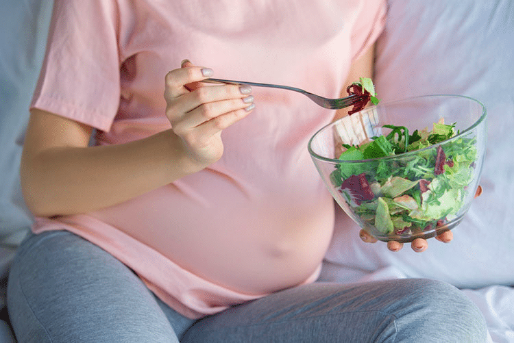 Pregnant woman eating salad sitting on the bed