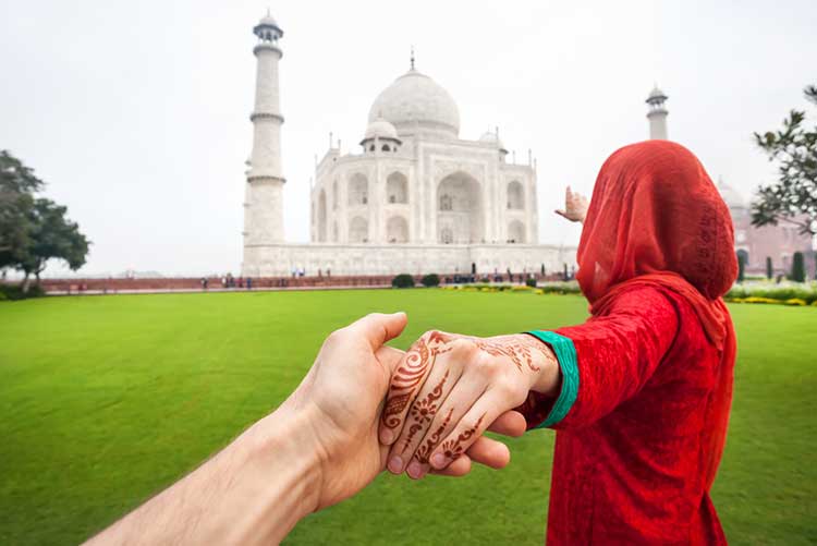 Woman in a traditional red salwar suit, holding her husband’s hand, pointing at Taj Mahal.